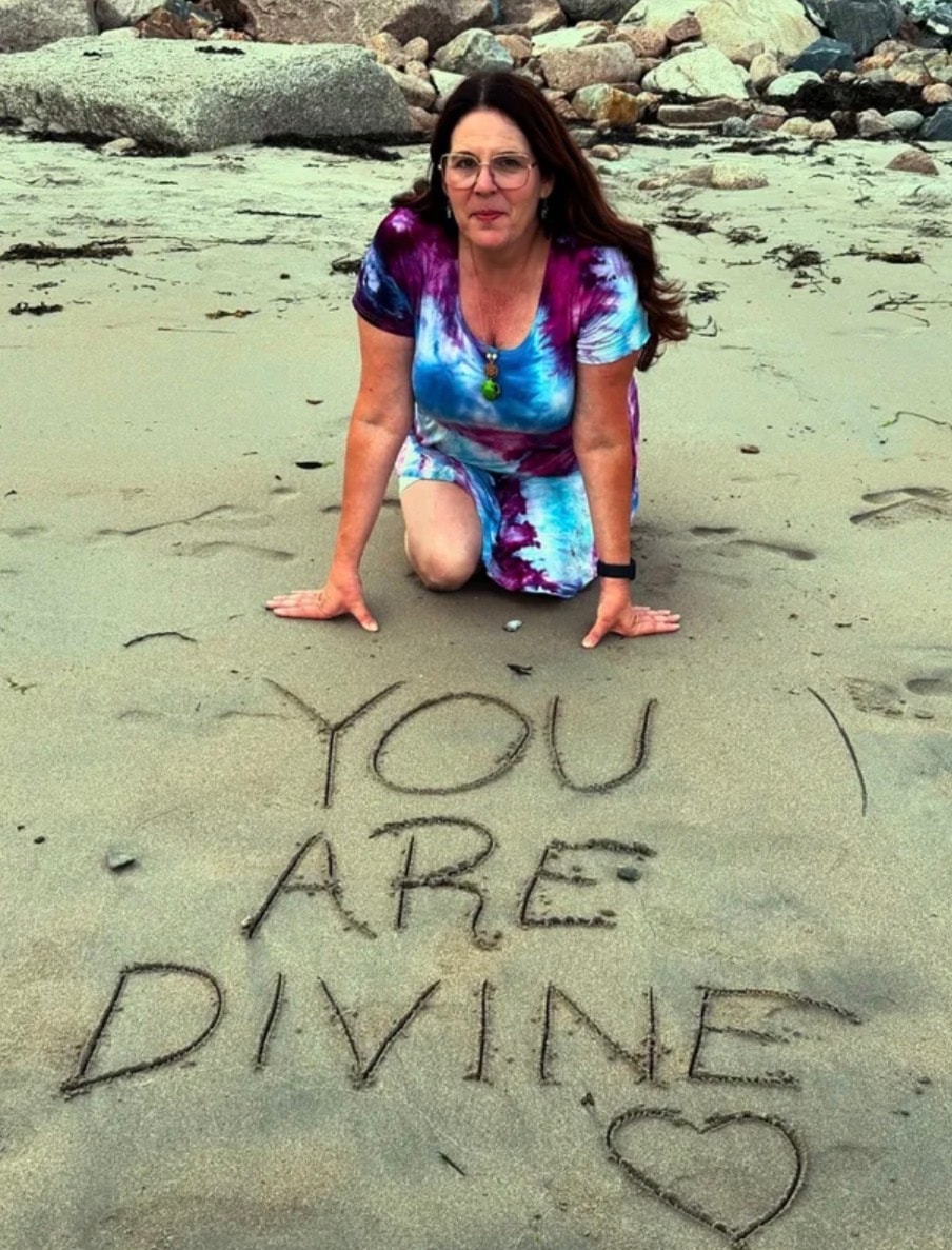 Angela kneeling on the beach beside You Are Divine written in the sand