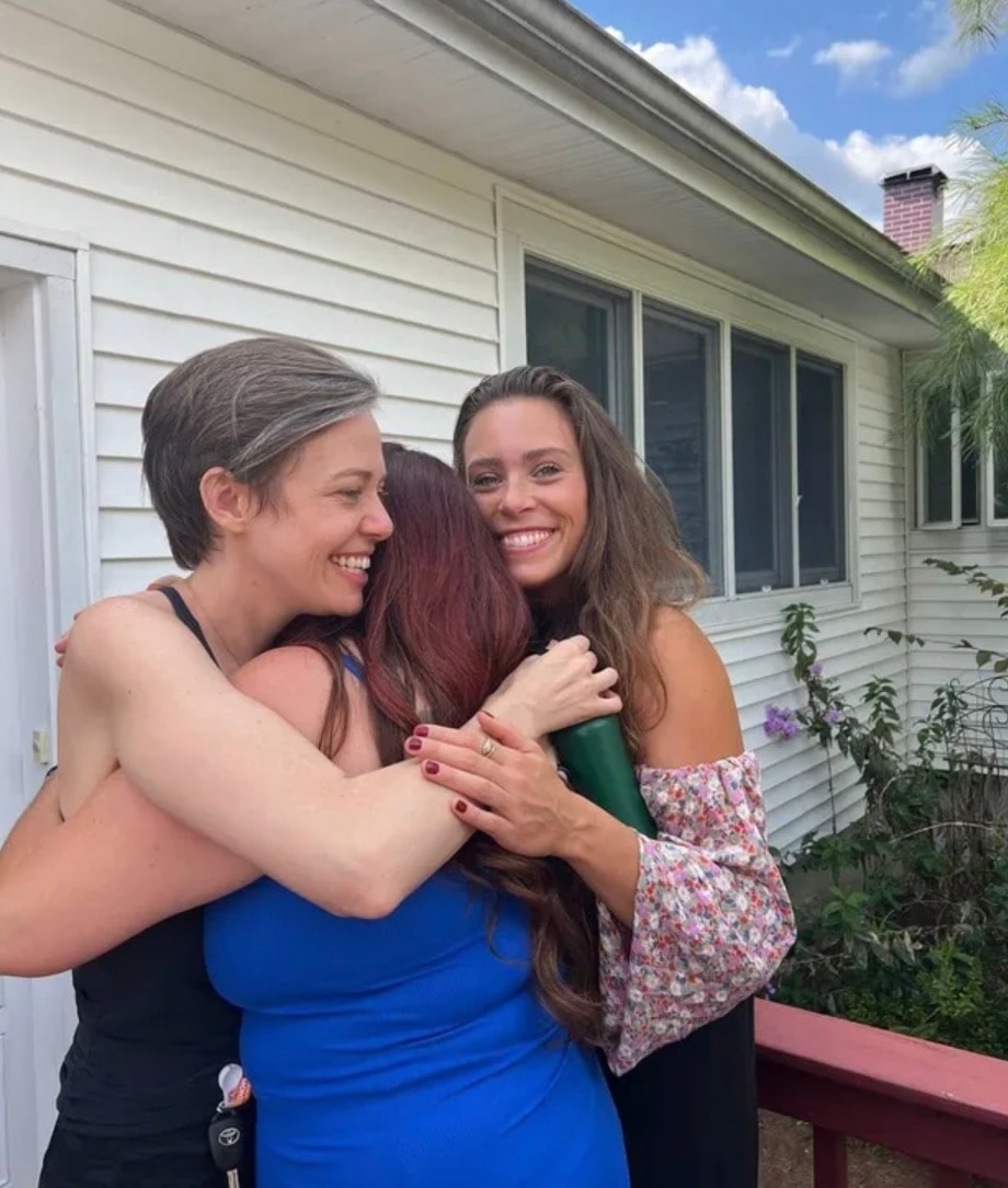 Three women sharing a warm group hug outdoors on a sunny day