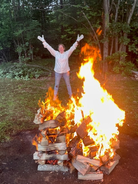 Angela standing triumphantly with arms raised behind a large bonfire