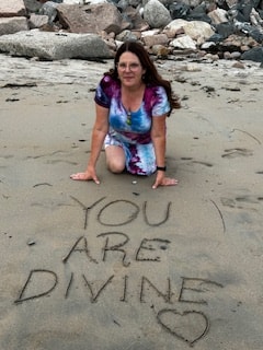 Angela kneeling on the beach beside 'You Are Divine' written in the sand