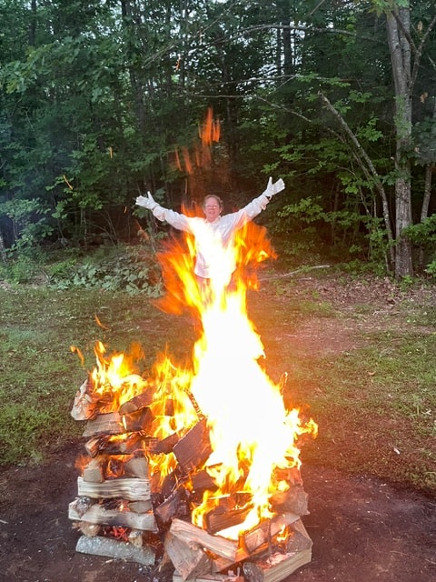 Angela with arms raised triumphantly in front of a blazing bonfire in a forest clearing